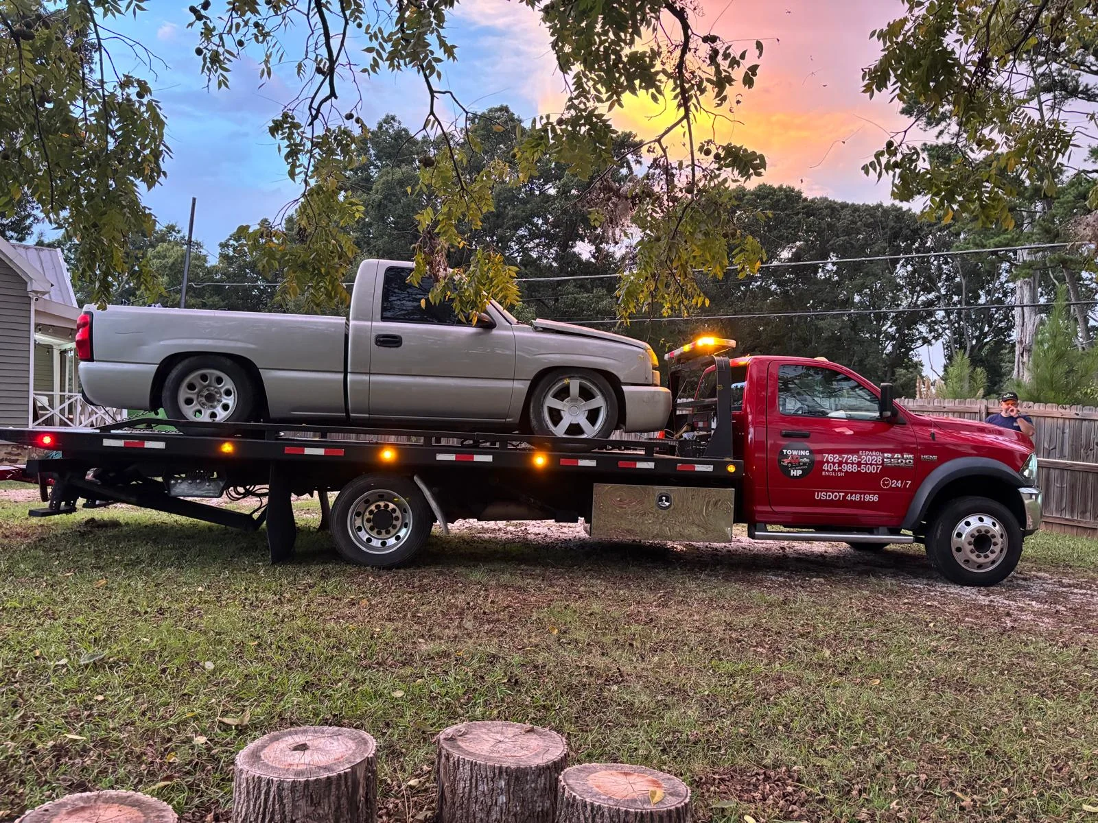 Red HP flatbed tow truck carrying a silver pickup at sunset